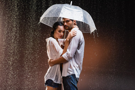 young couple in wet shirts standing under rain with transparent umbrella on dark backgroundの写真素材