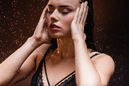 young woman in black lace bra posing under rain with closed eyes on dark backgroundの写真素材