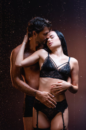 young man hugging seductive woman in black lace lingerie under rain on dark backgroundの写真素材