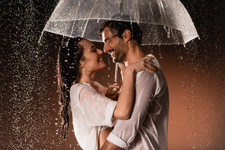 side view of happy couple embracing while standing with umbrella under rain on dark backgroundの写真素材