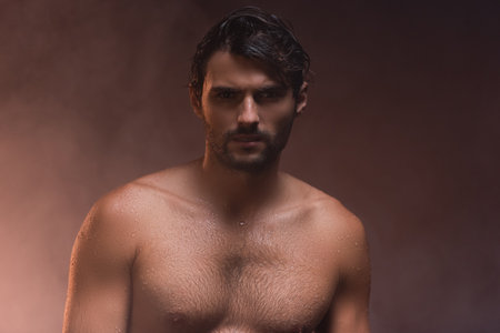 wet, shirtless man looking at camera while posing on dark background with smoke
