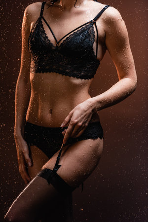 cropped view of sexy woman in black lace lingerie touching suspender belt under falling rain on dark backgroundの写真素材