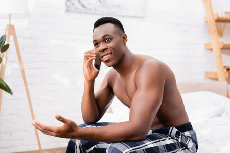 Shirtless african american man smiling while talking on smartphone in bedroomの写真素材