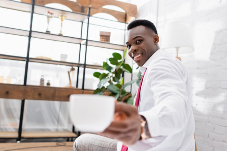 Cheerful african american businessman holding cup on blurred foreground at homeの写真素材