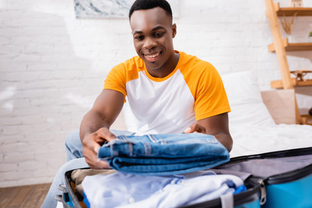 Smiling african american man putting jeans in suitcase on blurred foreground at homeの写真素材
