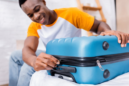 African american man locking suitcase on bed on blurred background