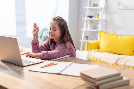 Smiling girl with waving hand looking at laptop at desk in living room on blurred foregroundの写真素材