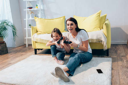Full length of excited mother and daughter with open mouths playing with gamepads on floor at home