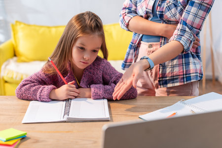 Upset daughter with pencil and notebook near mother pointing with hand near desk on blurred...
