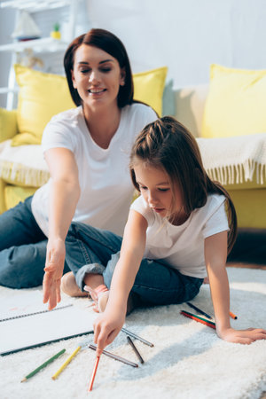 Daughter with colored pencil sitting near smiling mother on floor on blurred backgroundの写真素材