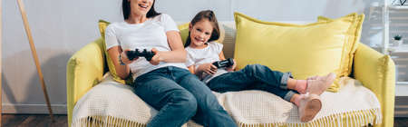 Happy daughter with joystick looking at camera near mother on couch, banner