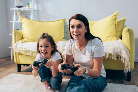 Happy mother and daughter playing with joysticks on floor on blurred backgroundの写真素材