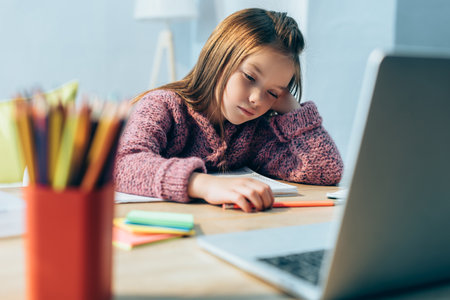 Sleepy girl at desk with stationery and blurred laptop on foreground