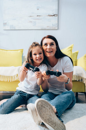 Happy mother and daughter playing with joysticks on floor on blurred foregroundの写真素材