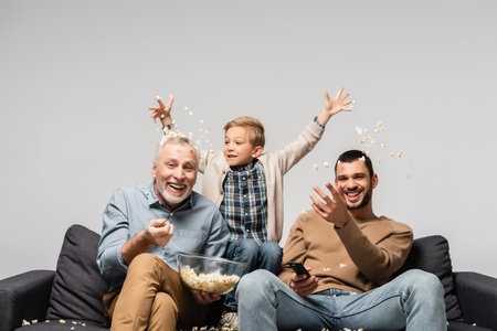 excited boy with dad and grandfather throwing popcorn while watching tv isolated on grayの写真素材