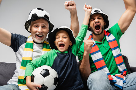 happy men and boy in fan hats shouting and showing win gesture while watching football match isolated on grayの写真素材