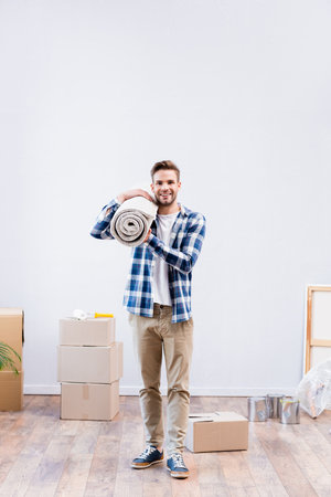 full length of happy young man looking at camera while holding carpet roll near cardboard boxes and paint tins at homeの写真素材