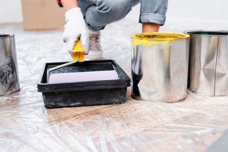 cropped view of young woman holding roller in tray near tins of paint on floor on blurred backgroundの写真素材