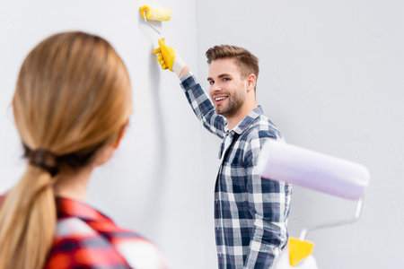 happy young man with roller painting wall while looking at blurred woman on foreground indoorsの写真素材