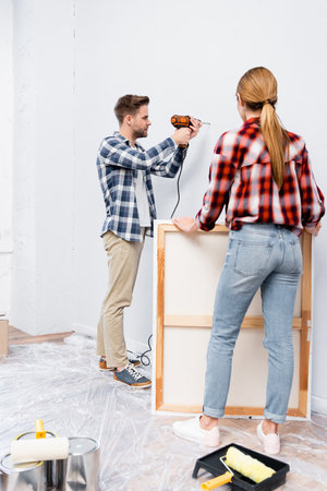 full length of young woman holding frame near man drilling wall at homeの写真素材