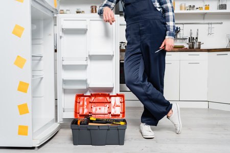 cropped view of young handyman with screwdriver leaning on fridge near toolbox in kitchenの写真素材