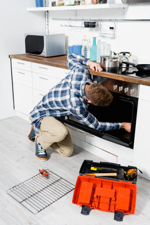 full length of young man repairing oven while sitting on floor near grid and toolbox in kitchenの写真素材