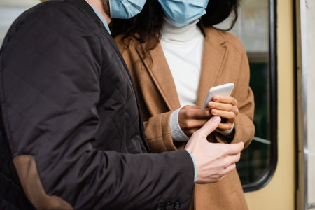 cropped view of african american woman in medical mask using smartphone near man in subwayの写真素材