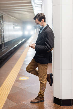 full length of man in medical mask standing and holding smartphone in hall of subwayの写真素材