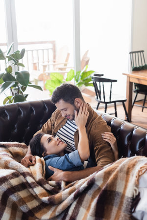happy young couple embracing while resting on leather couch under cozy plaid blanketの写真素材