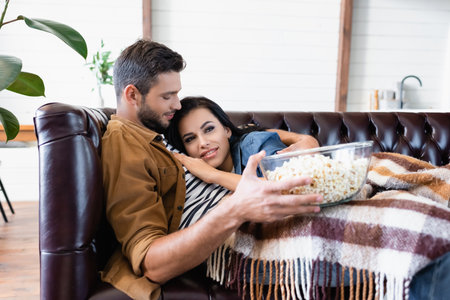young man holding bowl of popcorn while hugging girlfriend watching tv under plaid blanketの写真素材