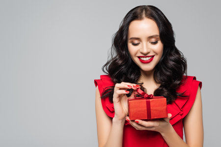 cheerful woman in ruffled dress pulling ribbon on gift box isolated on grayの写真素材