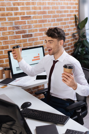 Positive forex businessman with takeaway coffee sitting near computers on blurred foregroundの写真素材