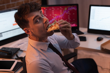 Businessman looking at camera while eating pizza in office at eveningの写真素材
