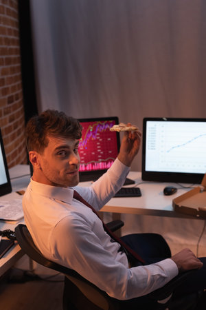 Businessman looking at camera while holding pizza near computers on blurred backgroundの写真素材