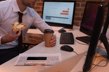 Cropped view of forex businessman holding delicious pizza and coffee to go near computers, news and smartphone on tableの写真素材