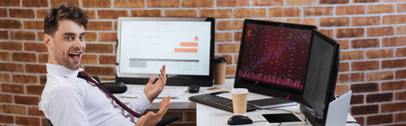 Positive forex businessman looking at camera near computers and coffee to go on table, banner