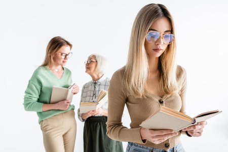 young woman in glasses reading book near mother and granny on blurred background isolated on whiteの写真素材