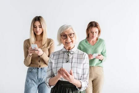 cheerful senior woman in glasses holding smartphone near daughter and granddaughter on blurred...
