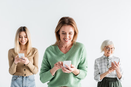 cheerful young woman using smartphone near mother and young daughter on blurred background isolated on whiteの写真素材
