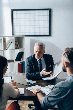 Smiling investor pointing with hand at paper folder near businesspeople on blurred foreground and laptop on tableの写真素材