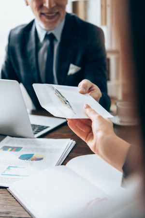 Cropped view of businesswoman taking envelope with money from investor near papers and laptop on...