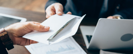 Cropped view of businessman taking envelope with money from investor near laptop on blurred...