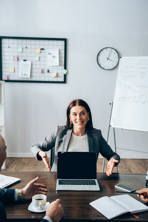 Businesswoman pointing at laptop near colleagues with coffee and notebook on blurred foreground