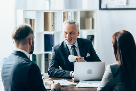 Smiling investor with cup of coffee looking at businesspeople on blurred foreground near papers and laptopの写真素材