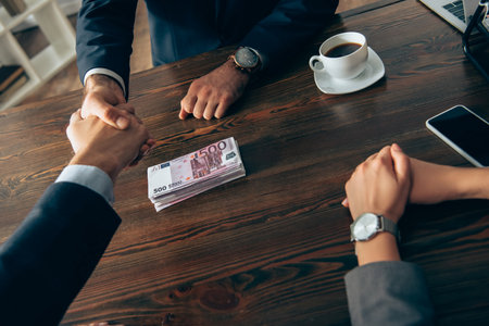Cropped view of investor and businessman handshaking near euro banknotes and businesswoman on blurred foregroundの写真素材