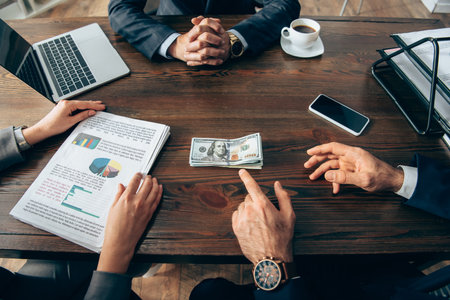 Cropped view of businessman pointing at money near colleague with papers and devices on table