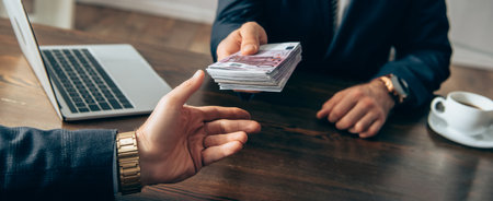 Cropped view of investor giving cash to businessman near laptop and coffee on blurred background,...