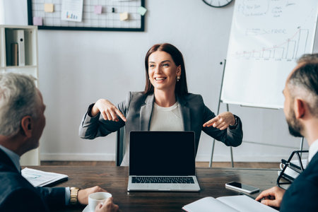 Smiling businesswoman pointing at laptop with blank screen near partners with notebook and coffee on...