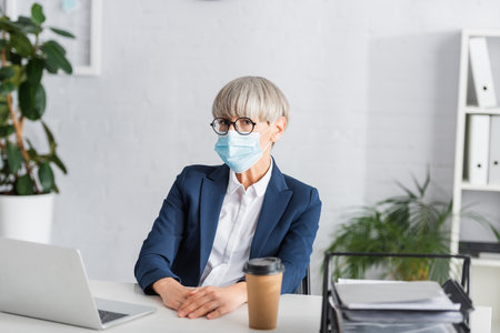 mature team leader in glasses and medical mask looking at camera near laptop and paper cup on deskの写真素材