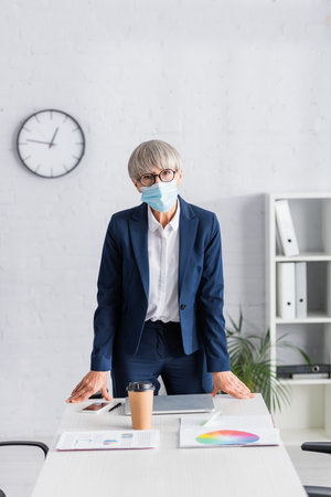 middle aged team leader in glasses and medical mask standing near workplace in modern officeの写真素材